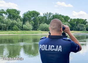 Policjant obserwuje jezioro przy pomocy lornetki.