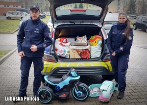 Policjant i policjantka stojący przy radiowozie wypełnionym prezentami świątecznymi.