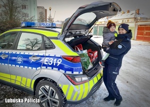 Policjantka trzyma na rękach małego chłopca, stojąc przy radiowozie policyjnym.