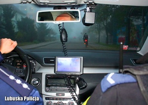 Policjanci jadący w radiowozie, a przed nim rowerzysta jadący drogą.